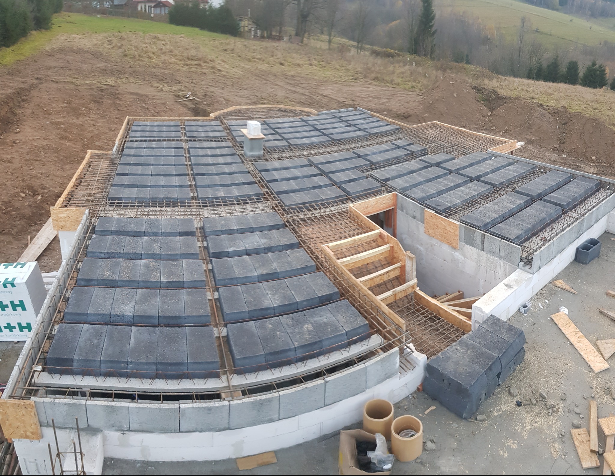 Widok z góry na szalunek fundamentów domu z ułożonymi bloczkami betonowymi i zbrojeniem, częściowo zalany betonem, z widocznym zejściem do piwnicy w trakcie budowy.