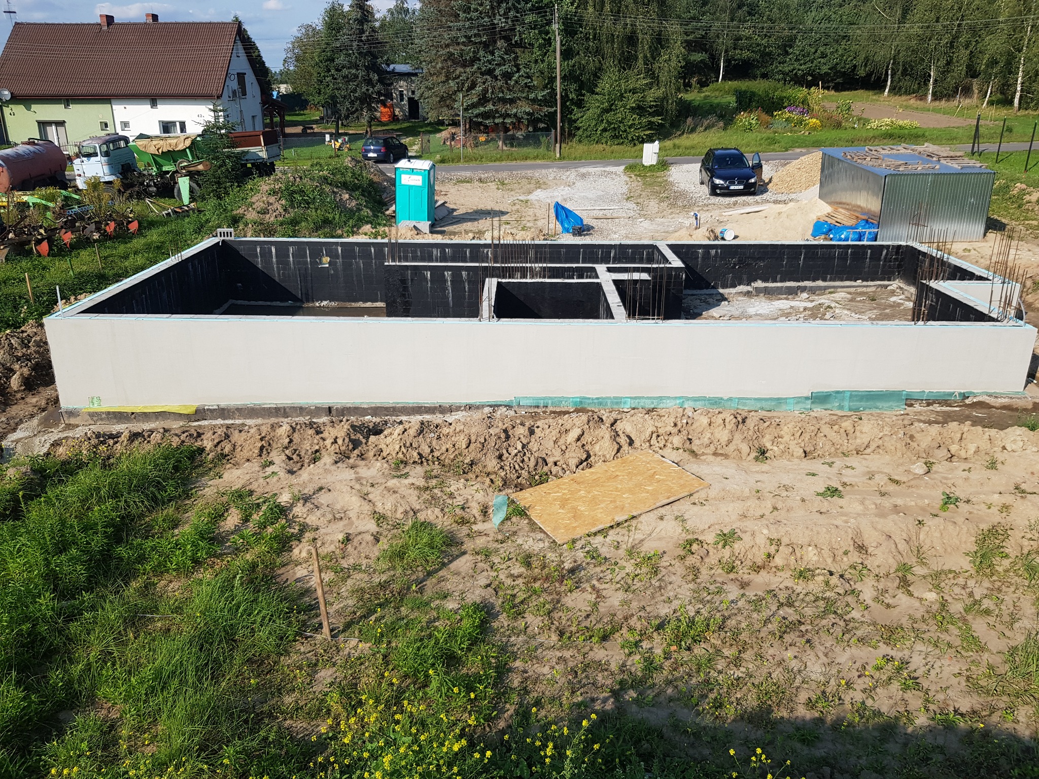 Wylane fundamenty domu jednorodzinnego z hydroizolacją, widoczne zbrojenia, szalunki i teren budowy z zapleczem socjalnym i materiałami budowlanymi.