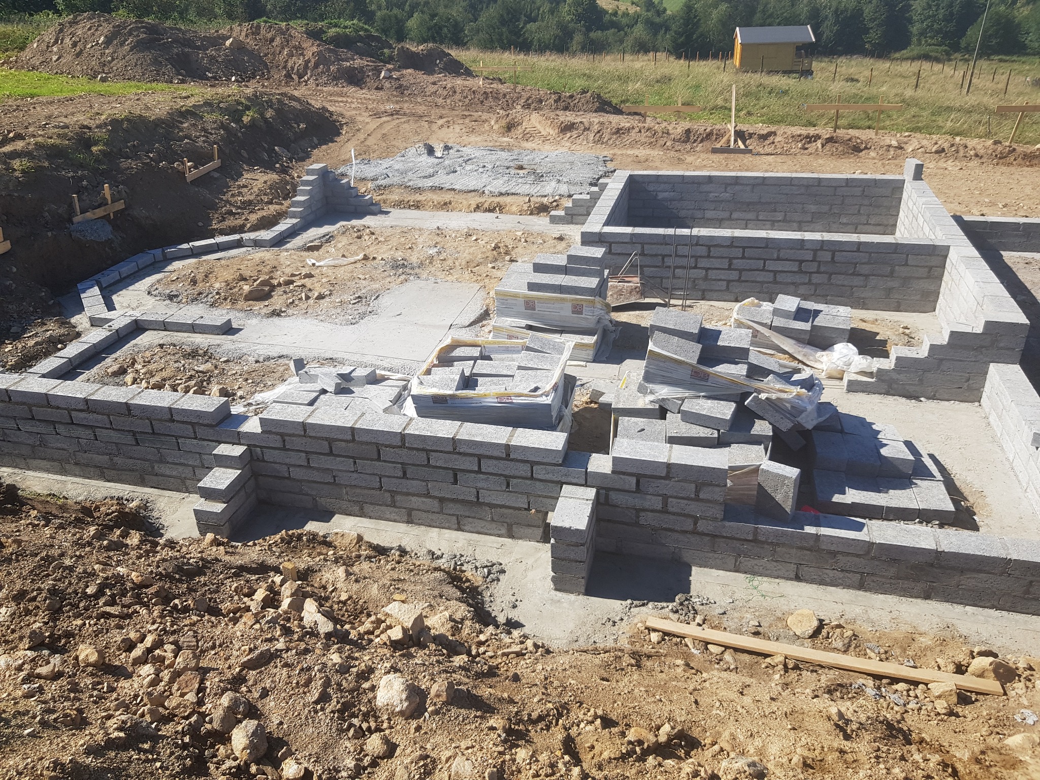 Wykop pod fundamenty z częściowo wymurowanymi ścianami z szarej kostki betonowej, widoczne palety z kostką i przygotowany teren, na tle zieleni i małego budynku.