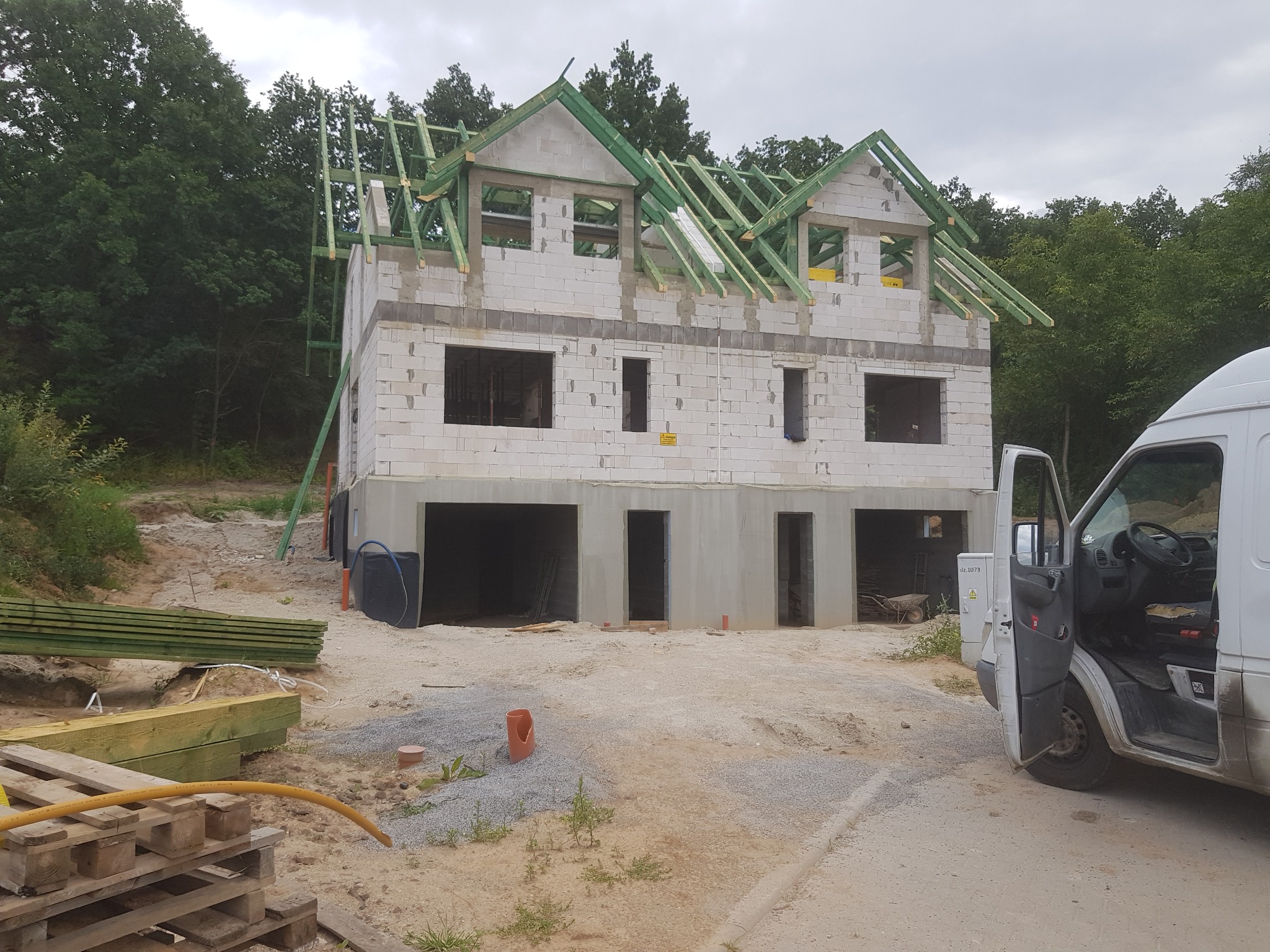 Dwukondygnacyjny dom w stanie surowym otwartym, z widoczną konstrukcją dachu z zielonych belek, białe bloczki, na pierwszym planie zaparkowany biały samochód dostawczy z otwartymi drzwiami, teren...