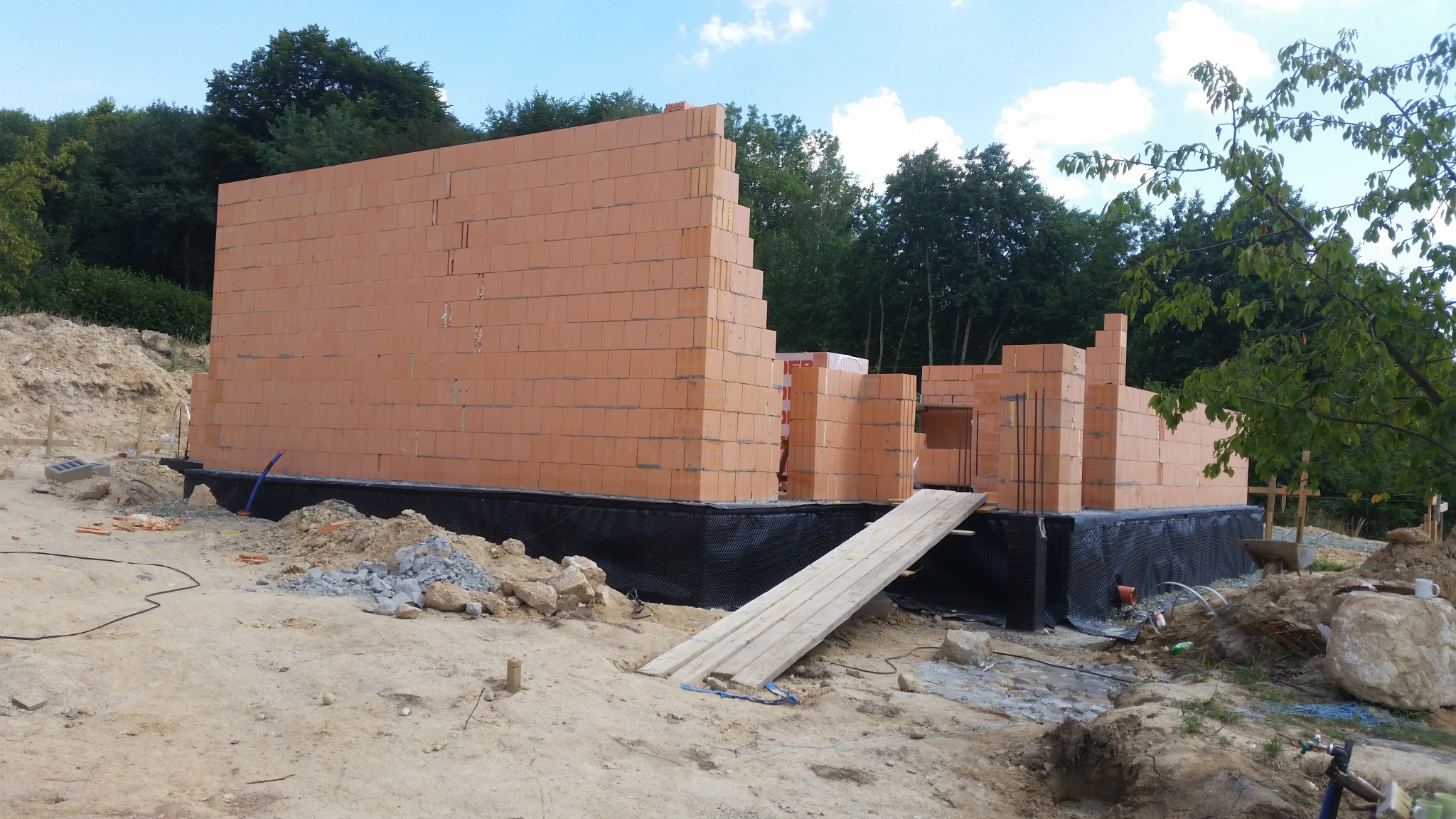 Wznoszone ściany parterowe domu z czerwonej cegły ceramicznej, widoczne zbrojenia i hydroizolacja fundamentów, teren budowy w trakcie prac.