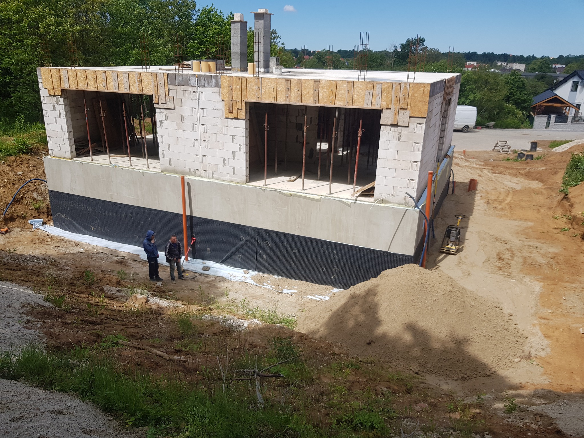 Wznoszenie ścian parteru domu z betonu komórkowego, widoczne otwory okienne z drewnianym szalunkiem, izolacja fundamentów oraz dwóch pracowników przy budynku w trakcie prac ziemnych.