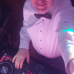 Uśmiechnięty DJ w białej koszuli i muszce, obsługujący mikser DJ-ski z podświetleniem w kolorze fioletowym.