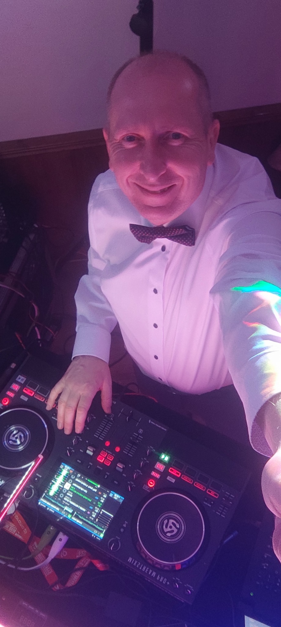 Uśmiechnięty DJ w białej koszuli i muszce, obsługujący mikser DJ-ski z podświetleniem w kolorze fioletowym.