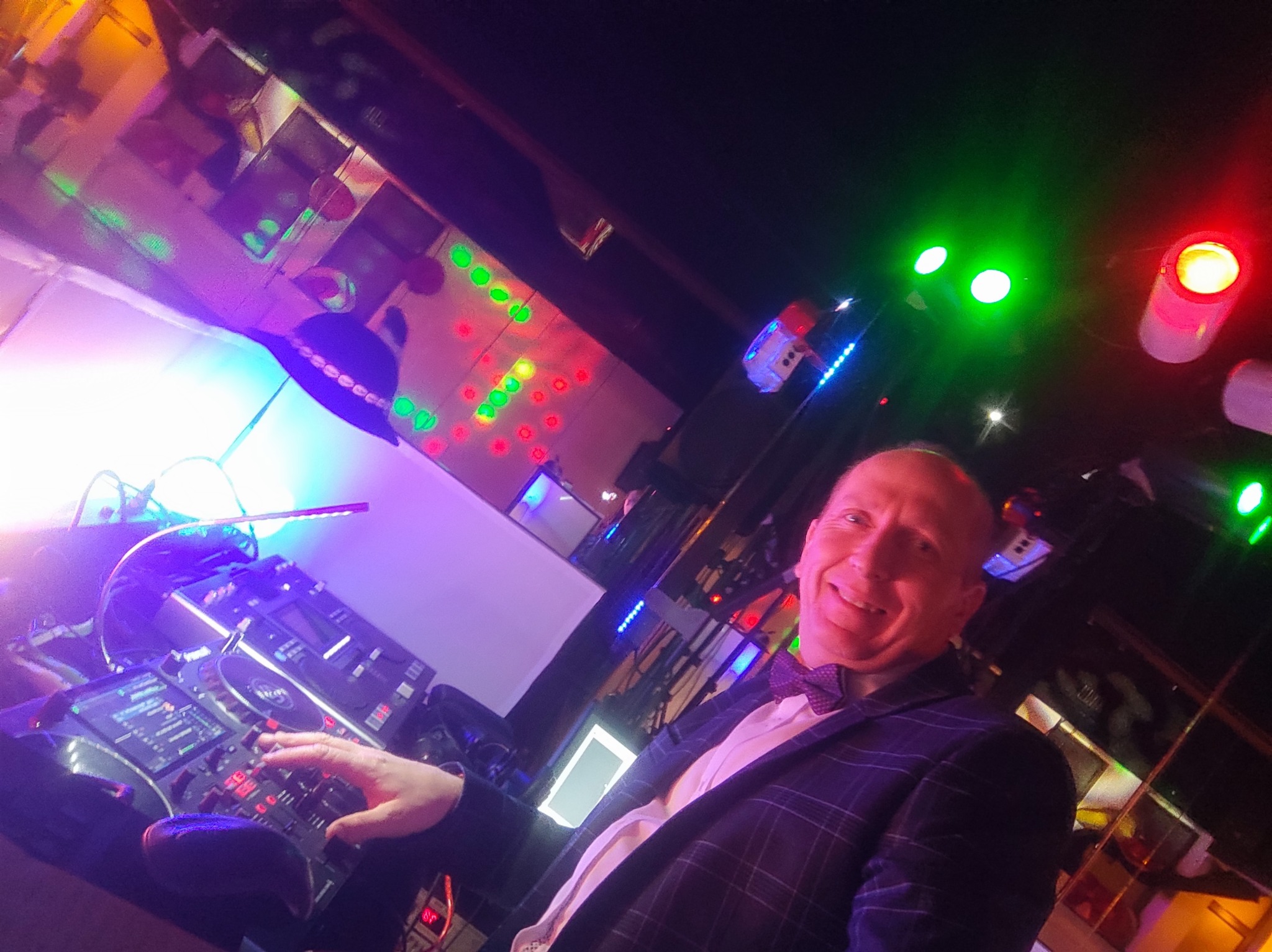 Uśmiechnięty DJ w muszce i marynarce, obsługujący konsoletę DJ-ską pod feerią kolorowych świateł na imprezie.