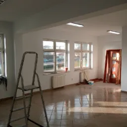 Wnętrze pokoju w trakcie odświeżania, widoczne świeżo pomalowane białe ściany, aluminiowa drabina, okna z widokiem na ulicę, grzejniki i drzwi z częściowo zdjętymi ościeżnicami.