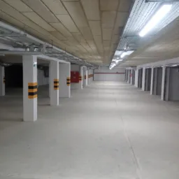 Wnętrze garażu podziemnego z betonową posadzką, rzędem białych kolumn z czarno-żółtymi pasami ostrzegawczymi, sufitem z paneli oraz oświetleniem w postaci lamp jarzeniowych.