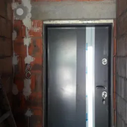 Nowe, czarne drzwi wejściowe z pionowym pasem ochronnym, zamontowane w surowym otworze muru z widocznymi przewodami elektrycznymi i śladami zaprawy.