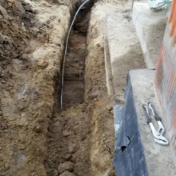 Wykop w ziemi z ułożoną rurą osłonową kabla, obok leżą kombinerki, a w tle widoczny młot udarowy na betonowej powierzchni.