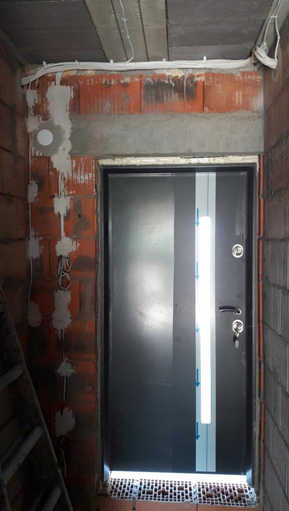 Nowe, czarne drzwi wejściowe z pionowym pasem ochronnym, zamontowane w surowym otworze muru z widocznymi przewodami elektrycznymi i śladami zaprawy.