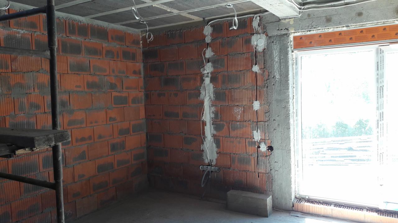 Wewnętrzna ściana z cegły z widocznymi przewodami elektrycznymi, częściowo zamurowanymi, surowy stan wykończenia, widoczne okno z jasnym światłem dziennym.