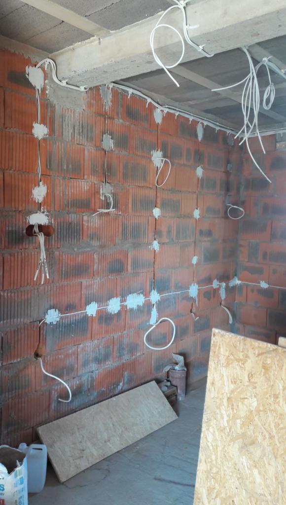 Wewnętrzna instalacja elektryczna w trakcie montażu w surowym budynku, przewody przymocowane do ściany z pustaków za pomocą gipsu, widoczne puszki instalacyjne i zwisające kable.