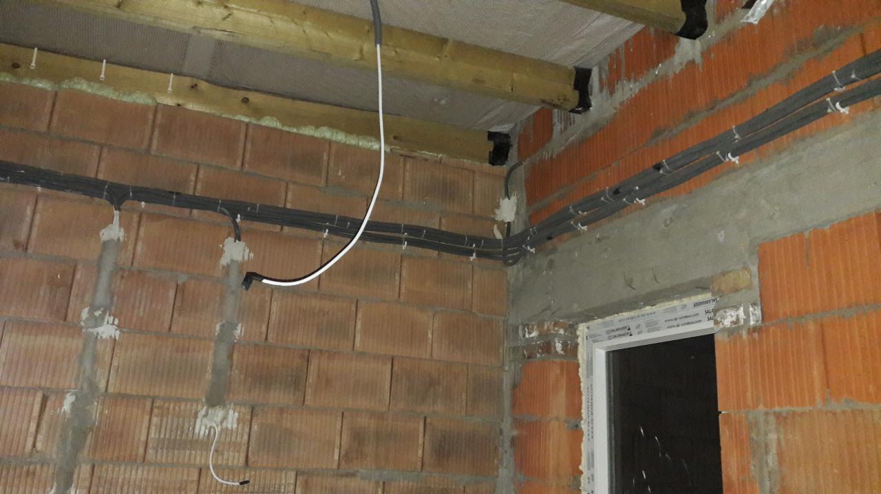 Surowy stan instalacji elektrycznej w budynku z czerwonej cegły, widoczne kable przymocowane do ściany i sufitu, fragment okna w prawym dolnym rogu.