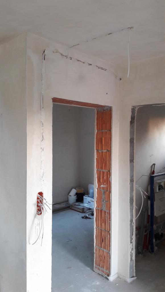 Wnętrze w trakcie remontu: widoczne przewody elektryczne wystające ze ściany, bruzdy na kable w suficie, surowy mur z cegły dziurawki przy ościeżnicy, oraz narzędzia i materiały budowlane w tle.