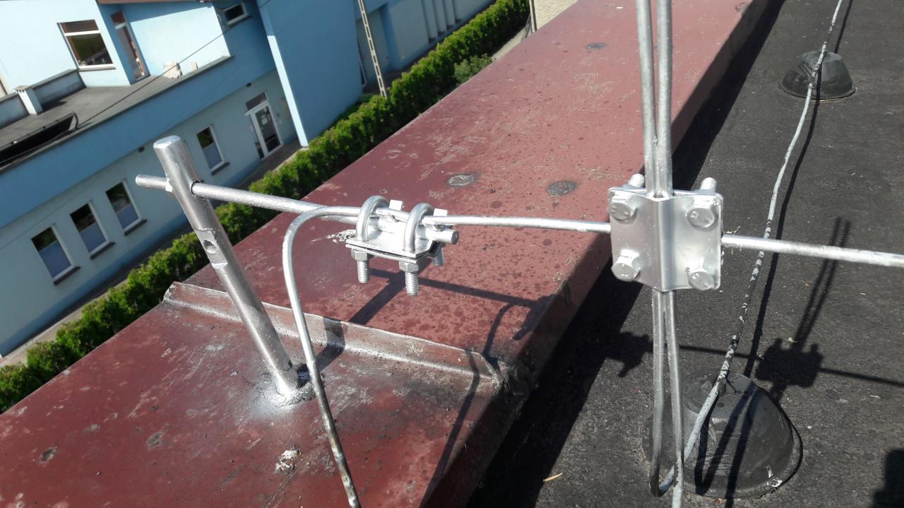 Metalowa instalacja odgromowa z mocowaniami na dachu budynku, widok z góry, w tle fragment budynku.