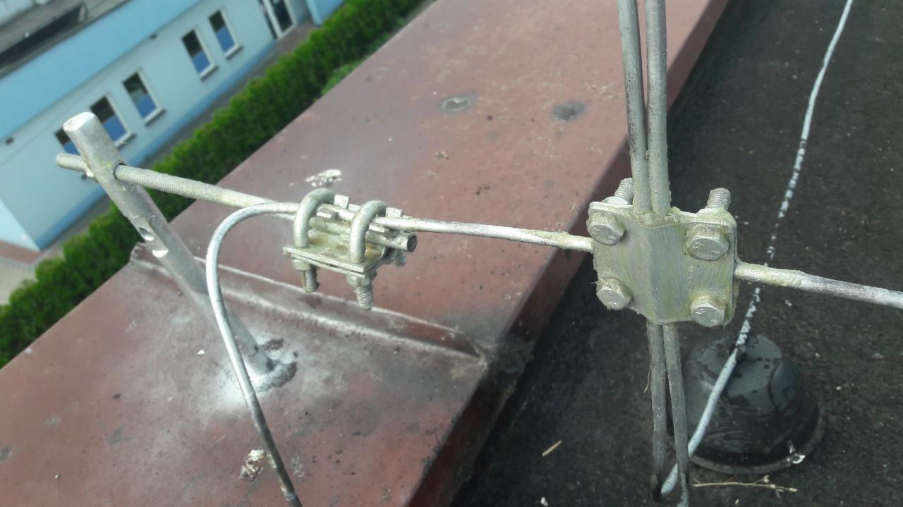 Metalowa konstrukcja odgromowa z mocowaniami na dachu budynku, widoczne ślady korozji i zabrudzenia, w tle budynek z zielenią.