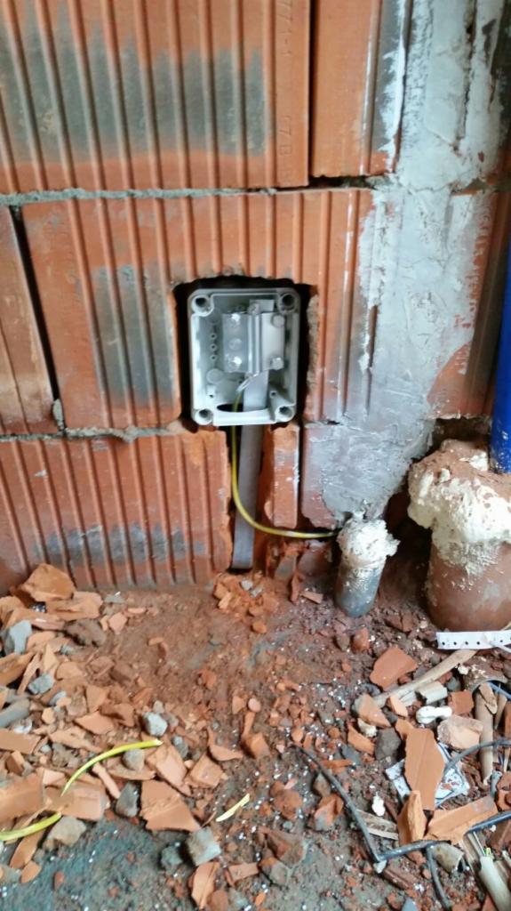 Montaż puszki elektrycznej w ścianie z czerwonej cegły, widoczny żółty kabel i fragment instalacji, gruz na podłodze.