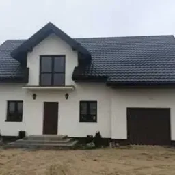 Nowoczesny dom jednorodzinny z ciemnym dachem z blachy na rąbek stojący, białą elewacją, ciemnymi oknami i drzwiami, na niezagospodarowanej działce.