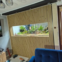 Krzysztof - Montaż drewnianych paneli ściennych z wbudowanym akwarium. Widoczne narzędzia i fragment niebieskiej sofy. Prace wykończeniowe w trakcie realizacji.