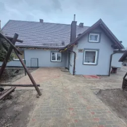 Szary dom jednorodzinny z nową elewacją i brukowanym podjazdem, oświetlony girlandami, z drewnianą huśtawką i studnią na pierwszym planie. Widok z poziomu gruntu.