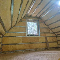 Poddasze z oknem w trakcie ocieplania: wełna mineralna między metalowymi profilami, widoczna konstrukcja dachu i drewniana podłoga. Etap prac przed wykończeniem.