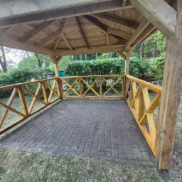 Drewniana altana z ażurową balustradą i brukowaną podłogą na tle zieleni. Widoczne elementy konstrukcyjne i detale wykończenia. Naturalne światło.