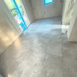 Szare płytki podłogowe ułożone w surowym wnętrzu z oknem zabezpieczonym folią. Widoczne przygotowane ściany do dalszych prac wykończeniowych. Minimalistyczny styl.