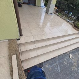 Krzysztof - Jasne płytki ceramiczne na schodach wejściowych domu, świeżo po ułożeniu. Widoczne fugi, fragment narzędzia i zabezpieczenie podłoża. Ujęcie z góry.