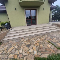 Krzysztof - Wykończone schody wejściowe do domu z jasnych płytek, z kamienną ścieżką prowadzącą do nich. Widoczna elewacja budynku w odcieniach zieleni i pracująca osoba.
