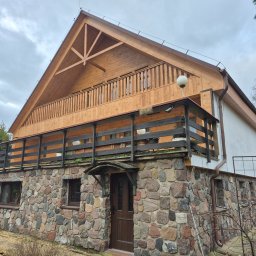 Krzysztof - Dwukondygnacyjny dom z kamienną elewacją i drewnianą górą, balkon z balustradą. Widoczne detale wykończenia i elementy otoczenia.