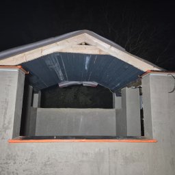 Krzysztof - Surowy stan elewacji budynku z drewnianą konstrukcją dachu, widoczne szare ściany i czarne elementy dachu w nocnym oświetleniu.