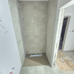 Krzysztof - Wnętrze łazienki w trakcie wykańczania: ułożone szare płytki w kabinie prysznicowej, widoczne instalacje wodne i fragmenty niezabezpieczonych ścian.