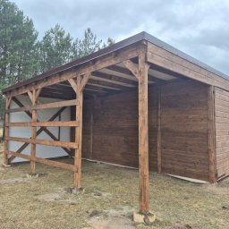 Krzysztof - Drewniana wiata z brązowym dachem na zewnątrz, z widoczną konstrukcją i białym materiałem w tle. Widoczne drzewa i trawa.