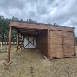 Krzysztof - Drewniana wiata z zamykaną częścią, wykonana z desek w odcieniach brązu, na tle drzew i pochmurnego nieba. Widoczna konstrukcja wsporcza i zadaszenie.