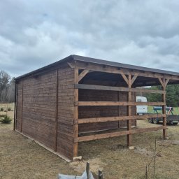 Krzysztof - Drewniana konstrukcja zadaszona brązowym blachodachówką, częściowo zabudowana deskami, na trawiastym terenie. W tle drzewa i pochmurne niebo.