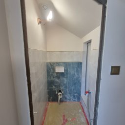 Krzysztof - Wnętrze łazienki w trakcie remontu. Widoczne płytki na ścianach, instalacja sanitarna i zabezpieczona podłoga. Oświetlenie punktowe i surowa rama drzwiowa.