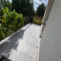 Krzysztof - Słoneczny balkon z jasnoszarymi płytkami gresowymi i ozdobną, ciemną balustradą, na której rośnie winorośl, otoczony zielonymi drzewami.