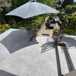 Krzysztof - Jasnoszary taras z płytek wielkoformatowych i metalowymi balustradami, prowadzący do zielonego ogrodu z dużym parasolem, meblami wypoczynkowymi i żółtymi kwiatami.
