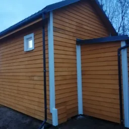 Nowoczesny, drewniany dom z elewacją w odcieniach brązu, z białymi elementami wykończeniowymi, takimi jak rynny i obramowania okien, sfotografowany o zmierzchu.