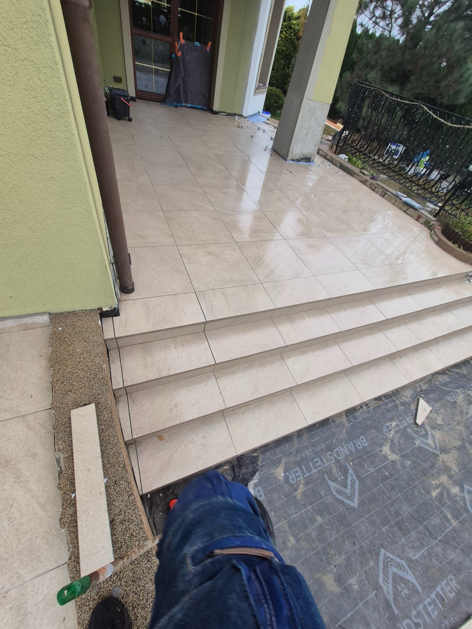 Jasne płytki ceramiczne na schodach wejściowych domu, świeżo po ułożeniu. Widoczne fugi, fragment narzędzia i zabezpieczenie podłoża. Ujęcie z góry.