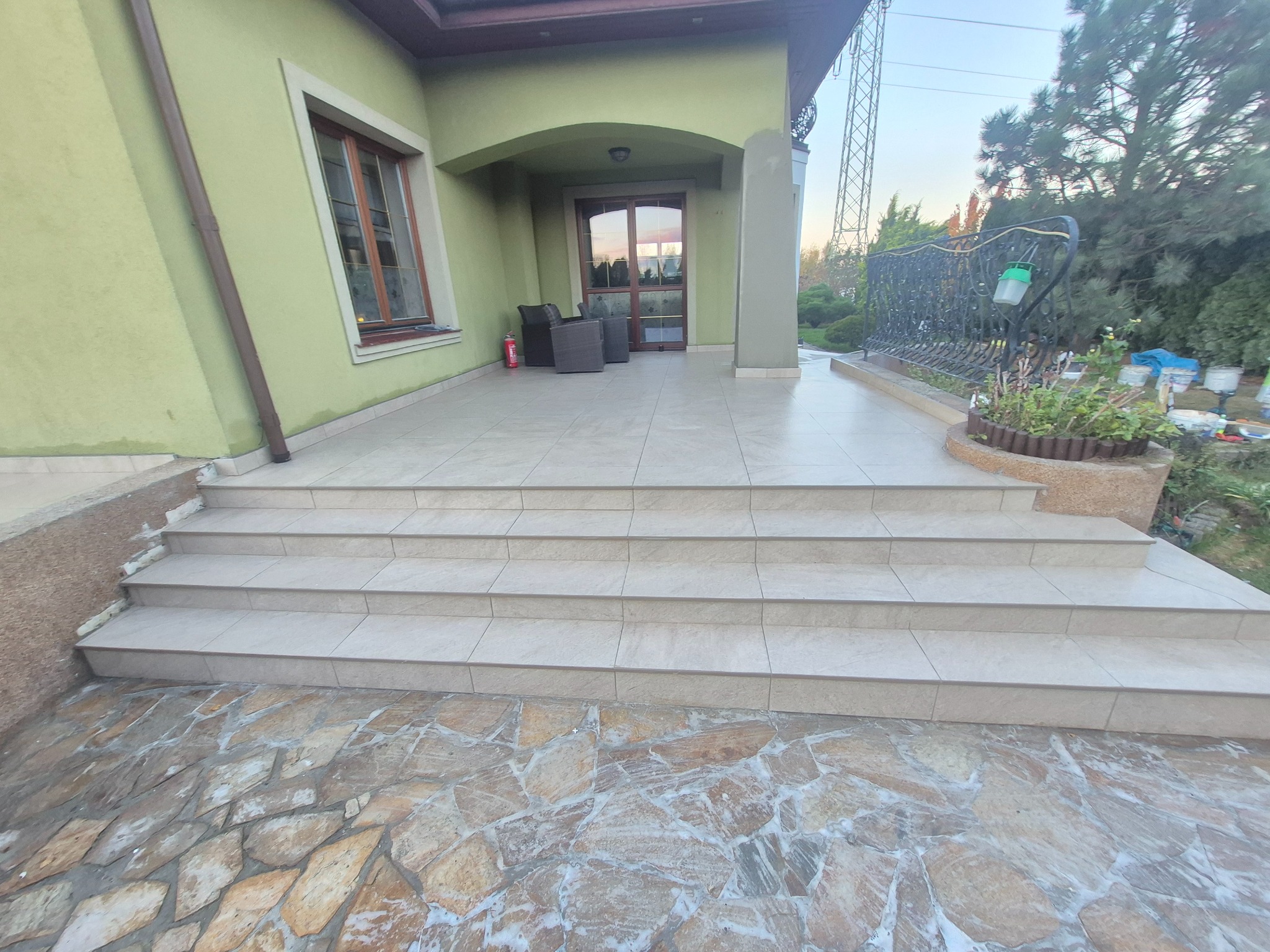 Wykończony taras z jasnymi płytkami i kamienną posadzką. Widoczne schody wejściowe i fragment elewacji domu w odcieniach zieleni. Na tarasie stoją meble ogrodowe.