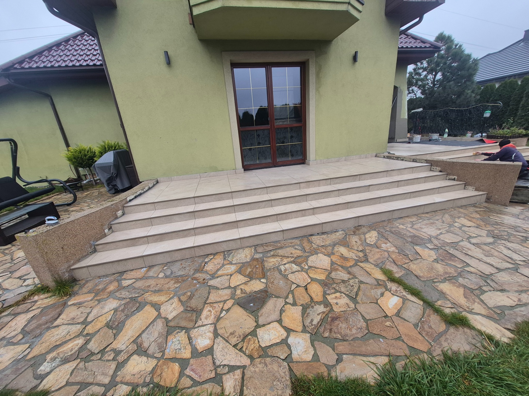 Wykończone schody wejściowe do domu z jasnych płytek, z kamienną ścieżką prowadzącą do nich. Widoczna elewacja budynku w odcieniach zieleni i pracująca osoba.