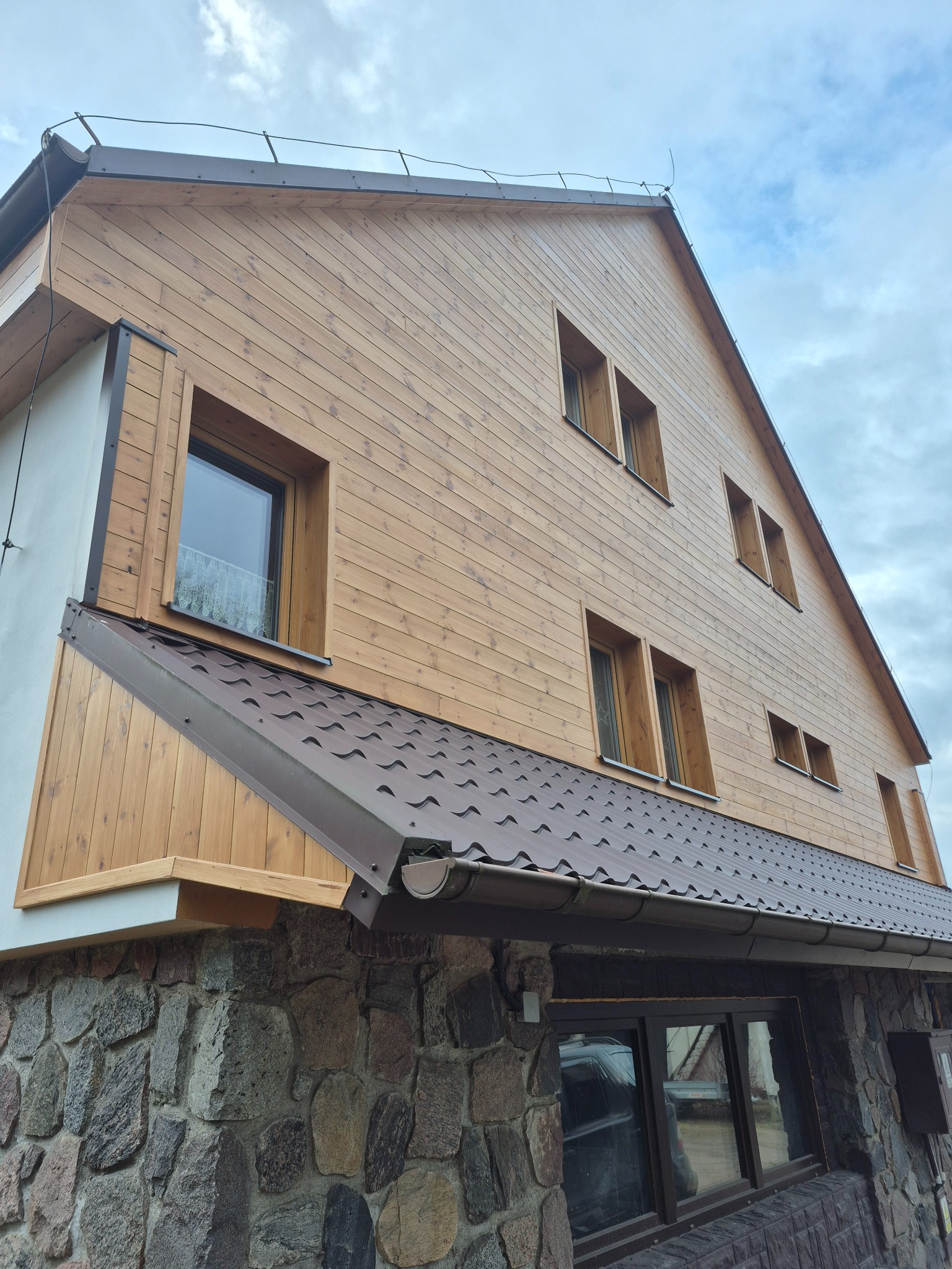 Wykończony dom szkieletowy z drewnianą elewacją i kamienną podmurówką. Widoczne okna z drewnianymi ramami i brązowy dach. Ujęcie z dołu, pochmurne niebo w tle.