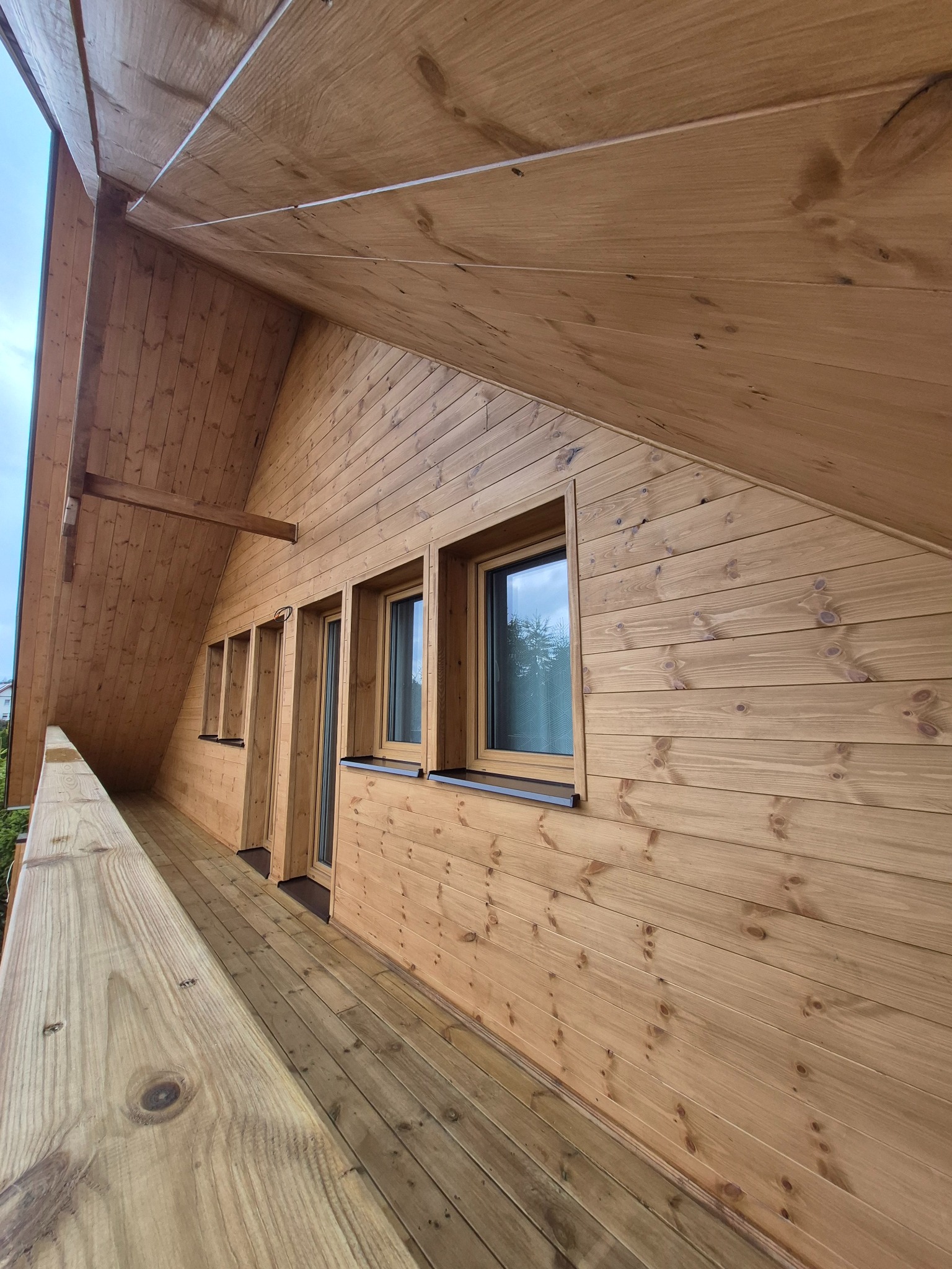 Drewniany dom szkieletowy z widocznym wykończeniem elewacji i balkonu. Naturalne drewno, okna z brązowymi ramami, skośny dach. Ujęcie z dołu, podkreślające strukturę drewna.