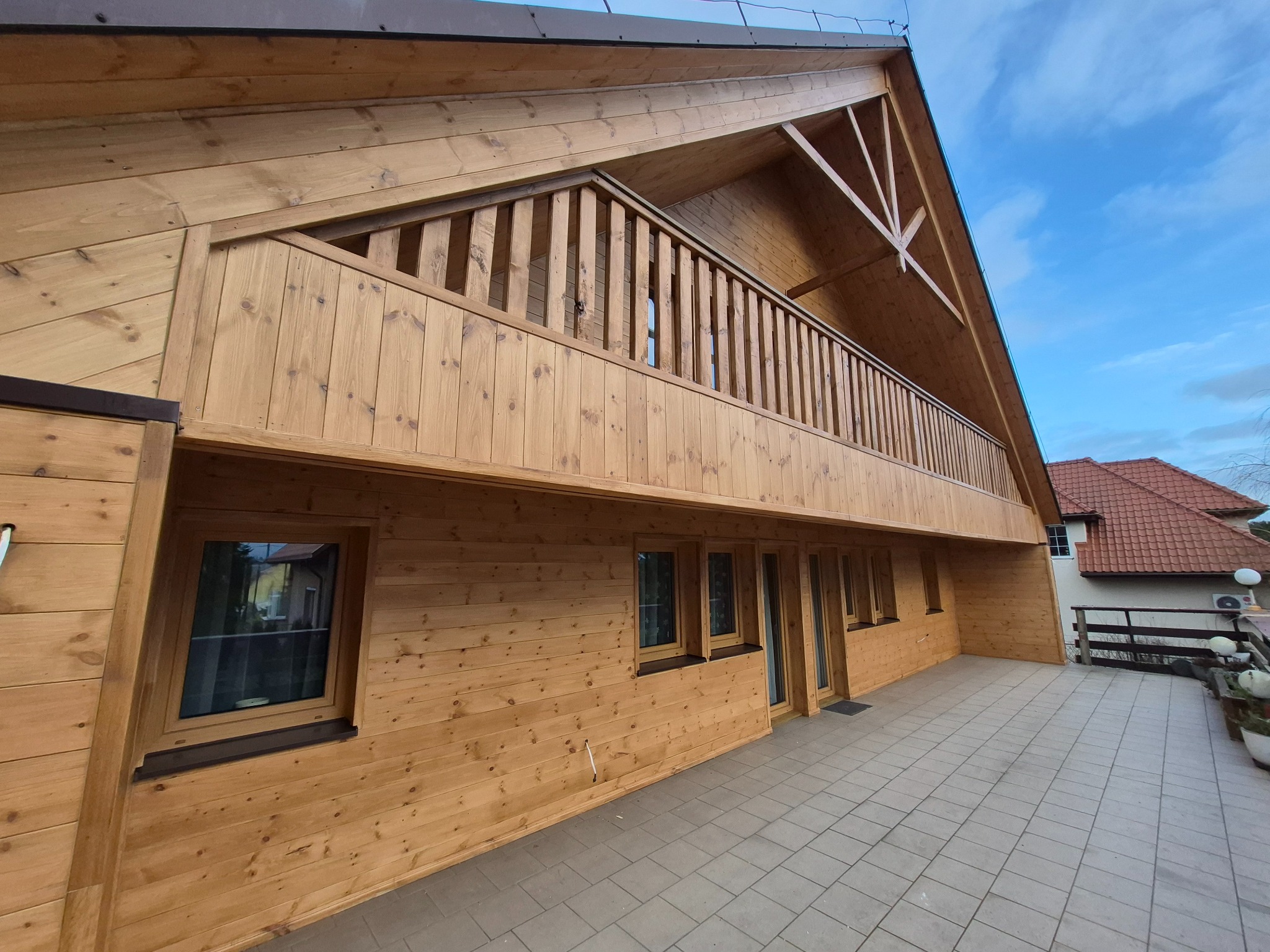 Jasny, drewniany dom szkieletowy z balkonem, widok z dołu. Charakterystyczna konstrukcja dachu i elewacji z desek. Szara kostka brukowa na tarasie.