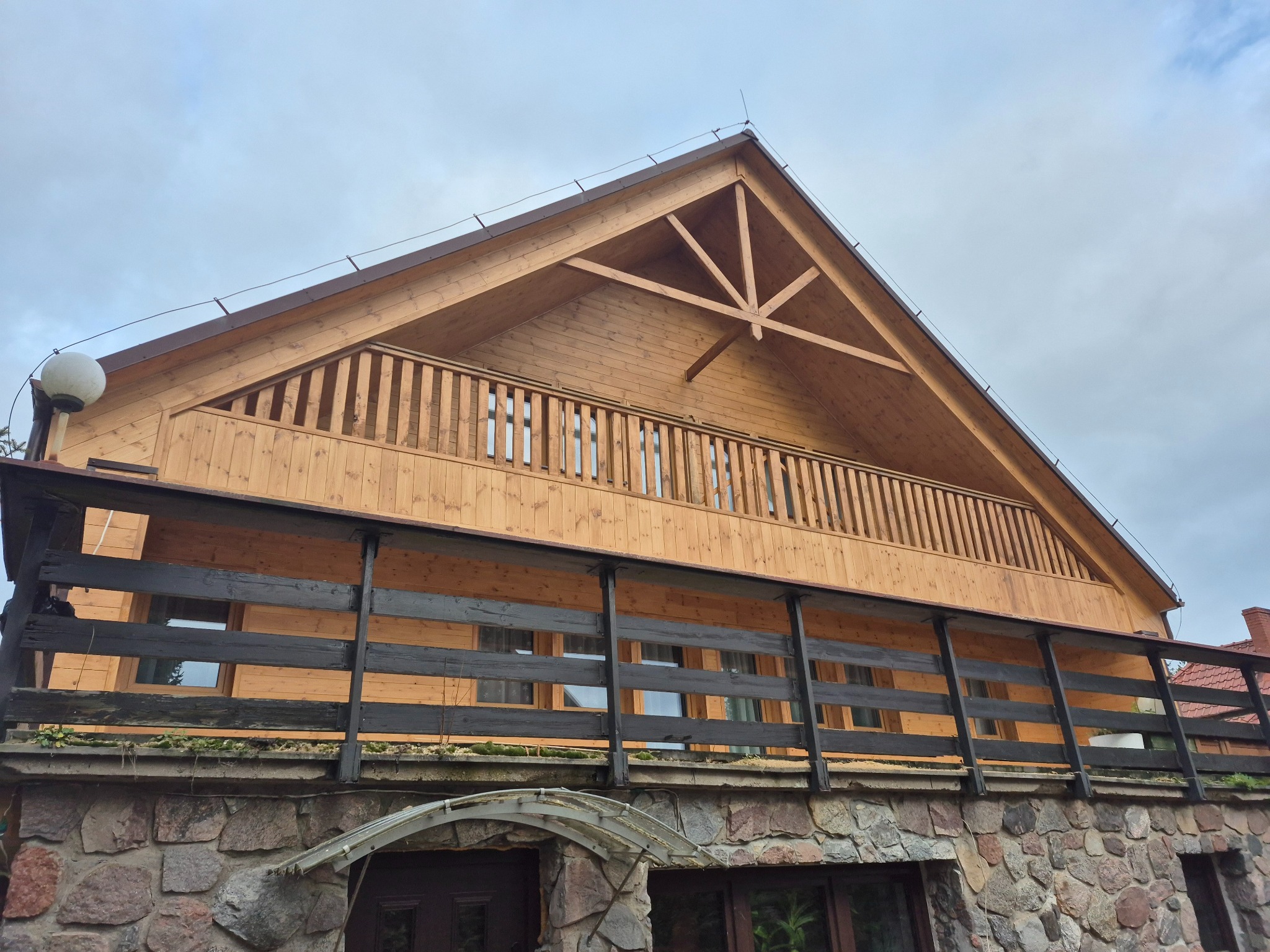 Dwukondygnacyjny dom z bali z balkonem i kamienną podmurówką, widok z dołu na drewnianą konstrukcję dachu i balustrady. Pochmurne niebo w tle.