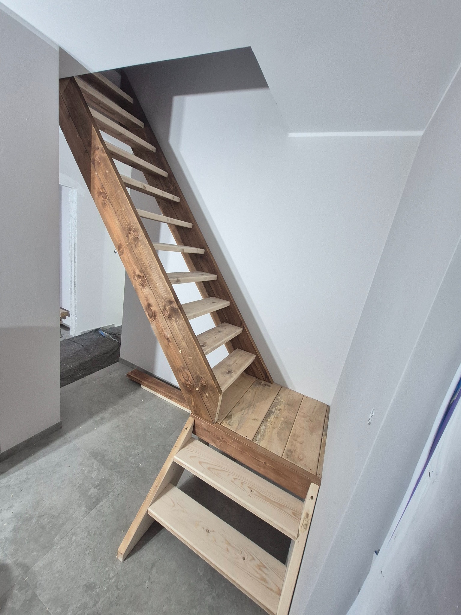 Drewniane schody strychowe z platformą, częściowo wykończone, z widoczną surową konstrukcją i fragmentem pomieszczenia w trakcie prac wykończeniowych.