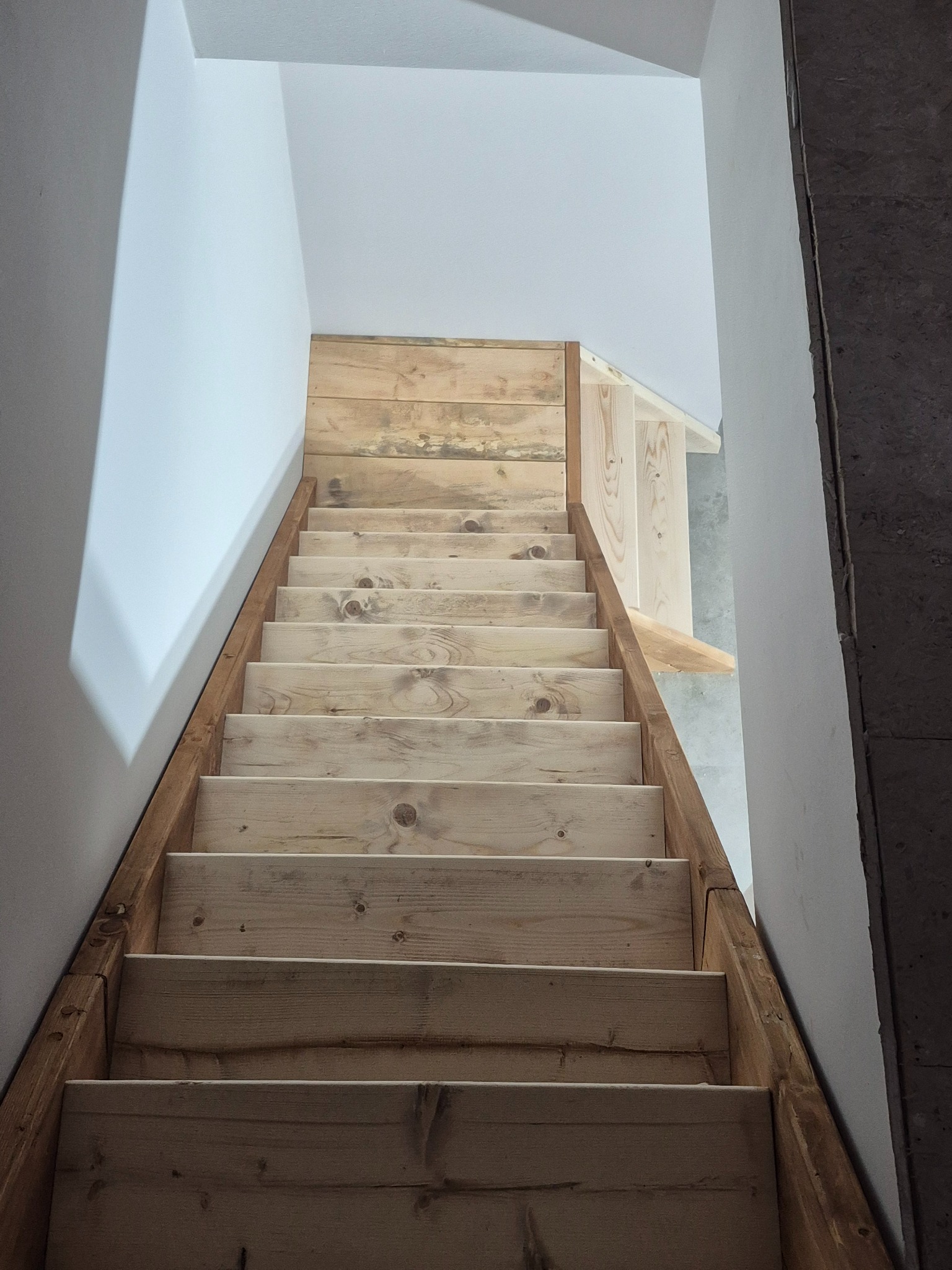 Drewniane schody w jasnym wnętrzu, widok z dołu. Naturalne drewno, proste wykończenie. Widoczne detale konstrukcyjne i surowe ściany. Ujęcie z perspektywy użytkownika.