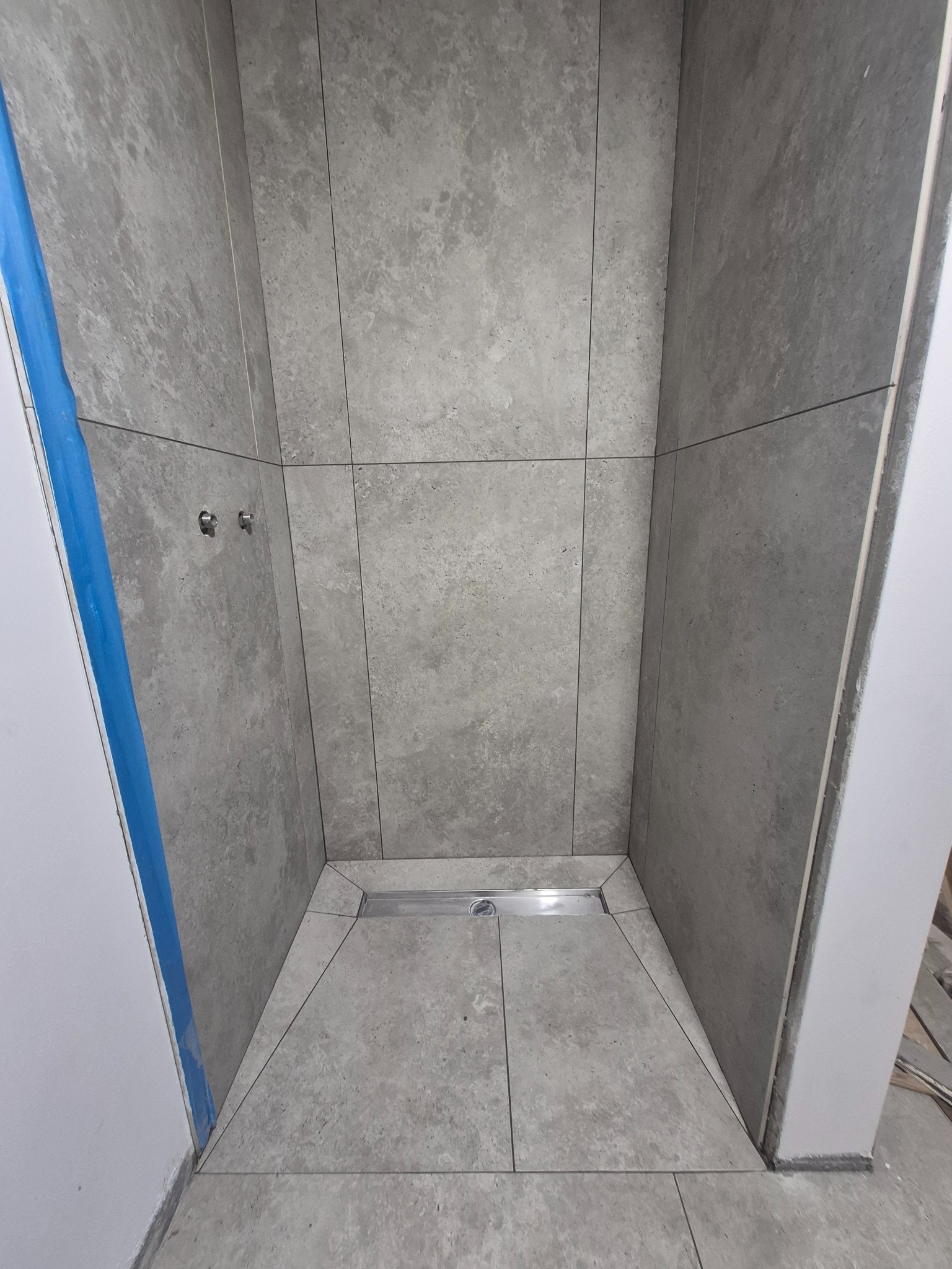 Wykończony brodzik z odpływem liniowym, wyłożony szarymi płytkami imitującymi kamień. Widoczne fugi i zabezpieczenia na ścianach. Minimalistyczny design.