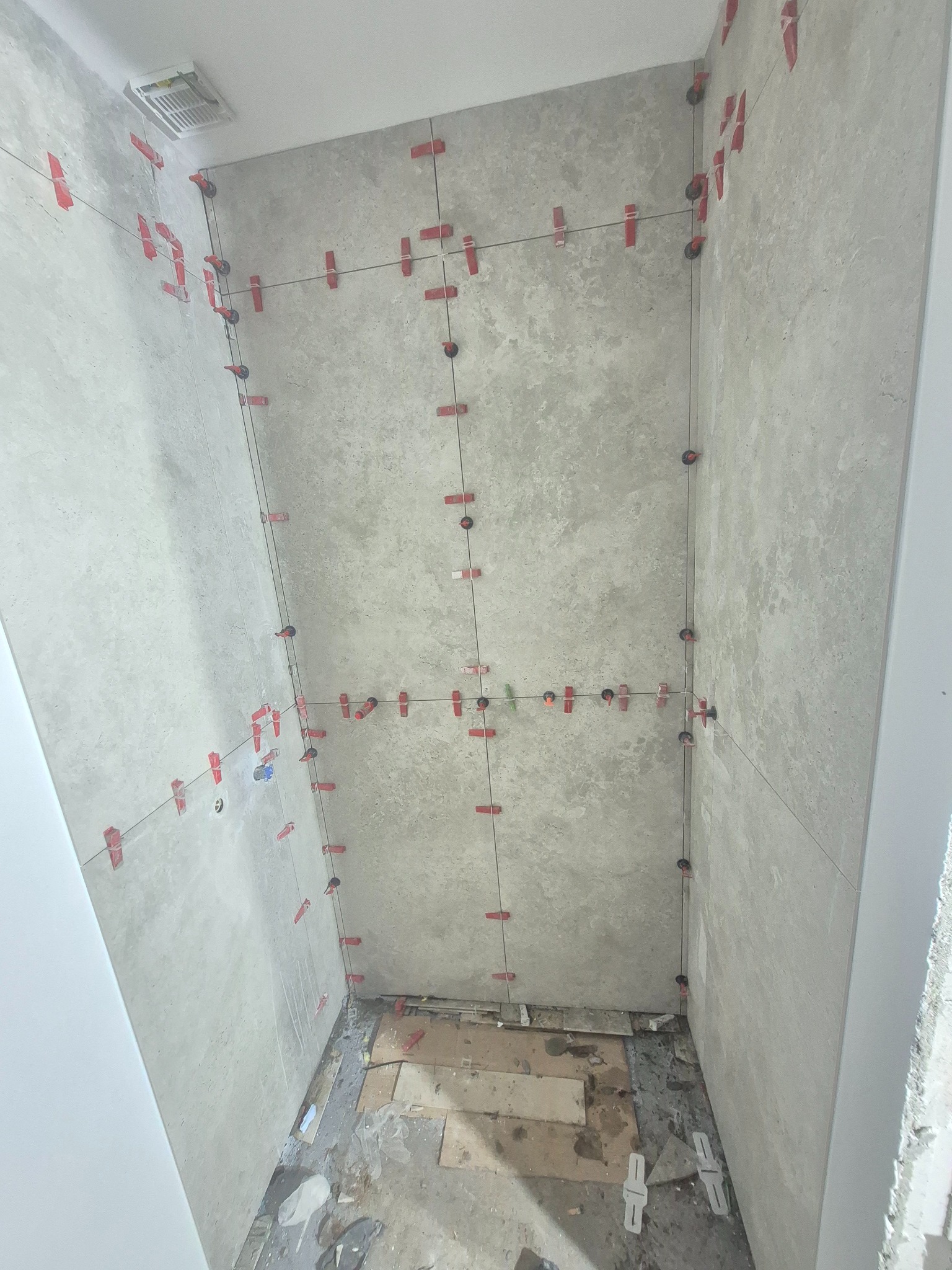 Ściany łazienki w trakcie układania płytek, z użyciem systemu poziomowania. Widoczne czerwone klipsy i kliny, szare płytki imitujące beton, surowy stan podłogi.